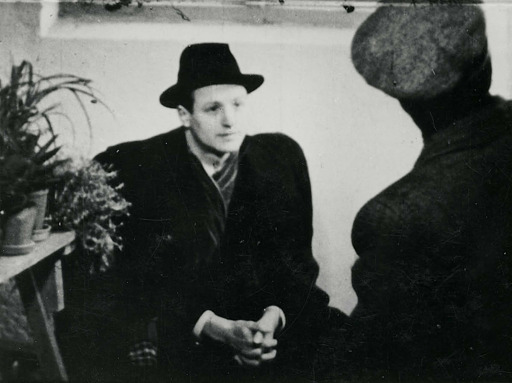 Still frame from the interrogation of Henning Walthing. Walthing is sitting down, wearing a fedora and an overcoat. To the right you can see a shelf with potted plants. Interrogation leader Andresen, wearing a sixpence, is standing at the right with his back against the camera.