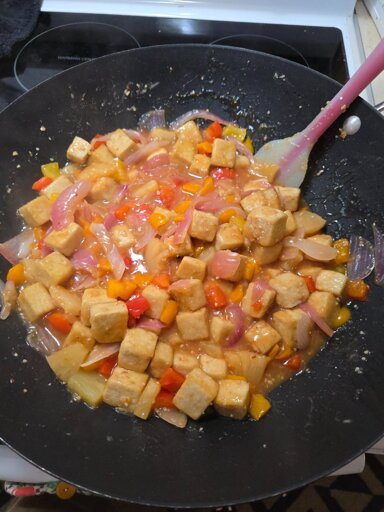 A wok with crispy cubed tofu, sliced red onion, bell pepper chunks,  pineapple chunks, a shit ton of minced garlic, shredded ginger, all covered in a creamy sweet and sour sauce. Some smoke emits from the pan.  